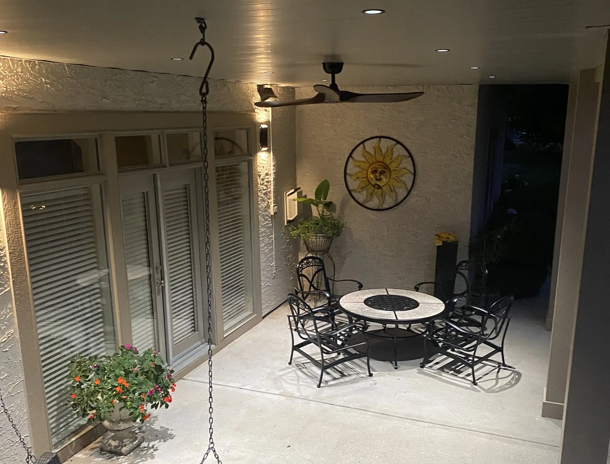 An under-deck patio at dusk, finished ceiling with recessed lights and a fan glowing over a stone dining table